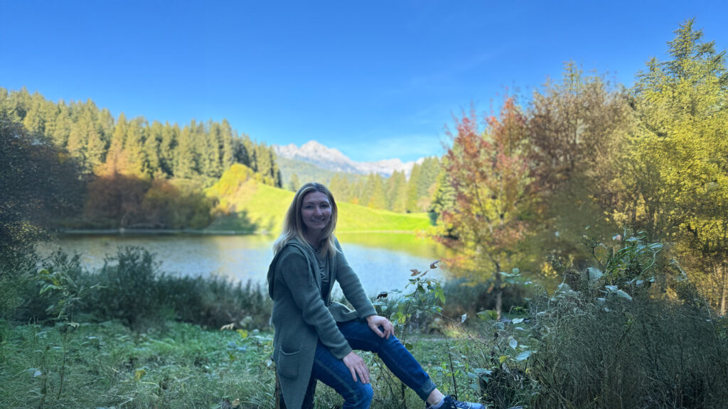 Petra sitzt am Ufer eines Sees mit Bergen im Hintergrund und blickt zur Seite