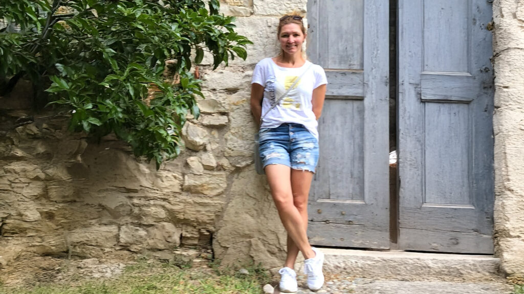 Petra steht entspannt in Shorts und T-Shirt vor einer alten Steintür und lächelt
